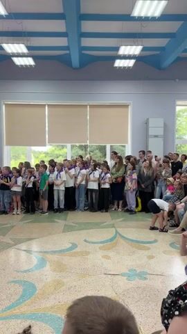 A group of children in white t-shirts and scarves are standing in a line indoors, facing a large screen. Behind them, a group of adults are also present. The room has a blue and white ceiling with lights, and large windows with blinds. The floor is decorated with a colorful pattern. The screen displays an image of children holding up signs. The video is shot from the perspective of someone in the audience, with the backs of their heads and shoulders visible in the foreground.