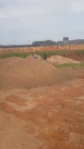 The media file shows an outdoor scene in Zawan, Nigeria. The primary subjects are a few individuals, possibly construction workers, engaged in an unspecified activity around large mounds of earth and sand. One person, dressed in dark clothing, is visible on a mound of soil. Another person, wearing lighter clothing, is also present. Scattered items, including a blue bag and some colorful debris, are visible near the mounds. The setting appears to be a construction or excavation site, characterized by piles of dirt and a cleared area. In the background, there are some buildings with dark roofs and a structure that resembles a water tower or industrial equipment. A green grassy strip runs along a fence or wall separating the construction area from the buildings. The ground in the foreground is a mix of reddish-brown earth and sand, with some patches of light-colored sand. The time of day appears to be overcast, with a bright but diffused sky suggesting it is daytime, possibly morning or afternoon. The weather seems to be mild and dry. There is no visible text in the media file. The overall impression is of a functional, work-in-progress environment.