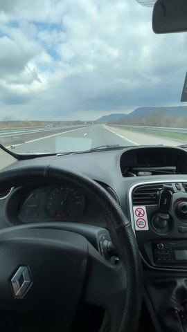 From the driver's seat, a vehicle travels on a multi-lane highway beneath a sky filled with puffy, white clouds that contrast with darker, stormy patches. Distant mountains are visible on the horizon. The vehicle's dashboard, steering wheel, and infotainment system are visible. A phone is mounted near the center console. There is a sticker indicating a maximum weight capacity of 490 kilograms for something. Other vehicles are seen traveling on the highway in the distance.