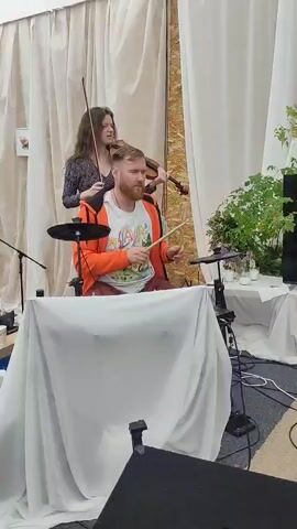 A group of musicians performs indoors. A woman in a dark floral dress plays the violin, standing to the left. A man in an orange hoodie and a white t-shirt sits behind a table draped with a white cloth, playing an electronic drum kit. Another man in a black t-shirt with white stripes stands behind him, appearing to be watching or possibly participating. The background is decorated with sheer white curtains and a potted plant. The lighting suggests an indoor venue.