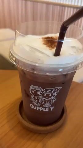 The video shows a close-up of a plastic cup filled with a chocolate drink. The cup has a clear plastic lid and a brown straw sticking out. The drink is topped with a thick layer of white foam, and there are some brown sprinkles on the foam. The cup is sitting on a small, round coaster on a wooden table. The cup has a logo on the front with the word "CUPPLEY" written below two cartoon figures. The background is a blur of cream and brown colors. The straw is being used to stir the drink.