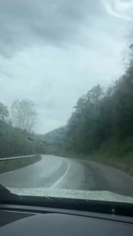 The view from inside a car shows a winding road through a hilly, forested area on a rainy day. The car's windshield wipers are on, clearing the rain from the glass. The road is wet and shiny, with white lane markings. The surrounding landscape is mostly green, with trees lining the roadside. The sky is overcast and gray.