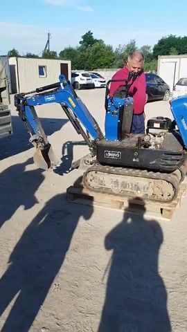 In the video, a man in a red shirt is standing next to a blue mini excavator. Another man in a grey t-shirt and denim shorts approaches the excavator from the right, carrying a blue plastic container. The man in the red shirt appears to be demonstrating or explaining something about the excavator to the man with the container. The scene takes place outdoors on a sandy surface with some buildings and parked cars in the background. The sun is shining, casting long shadows. The excavator is on a wooden pallet.