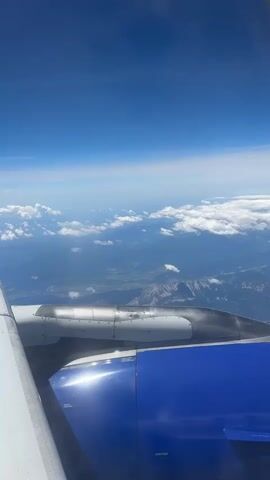 This is a first-person view from an airplane window, looking out at the sky and landscape below. The airplane's wing and engine, painted blue and gray, are prominently in the foreground. The sky is a vibrant blue with scattered white clouds. Below, the landscape appears mountainous and possibly rural, with patches of green and darker areas that could be forests or lakes. The sun is shining brightly, reflecting off the airplane's wing and engine.