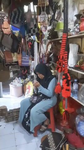 A woman wearing a blue hijab and a light blue dress sits on a red stool in what appears to be a shop. She is looking down and appears to be sewing or working with her hands on a piece of fabric. The shop is filled with various items, including bags hanging from the ceiling, shelves stocked with various objects like vases and what look like busts, and items wrapped in plastic. A decorative rope-like object with red and black pom-poms hangs prominently in the foreground. The lighting is somewhat dim.
