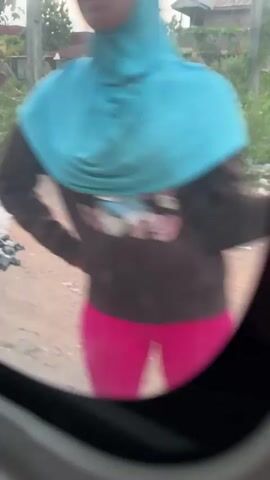 From inside a vehicle, the video shows a view through a window of a dirt road. A person wearing a purple shirt and patterned shorts walks on the side of the road. Vegetation grows along the roadside. Buildings are visible in the background.