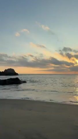 The video shows a beach at sunset.  The sky is a mix of light blue and orange, with clouds partially obscuring the sun.  The ocean water is calm with small waves. Two people are swimming in the water.  There are rocks on the left side of the frame.  The sand is wet near the water.