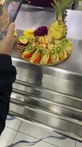 The video shows a fruit platter being prepared.  Two identical platters are visible, each featuring sliced kiwi, orange, apple, dragon fruit, mandarin orange, banana, and a pineapple cut to have a decorative top. The platters are displayed on a stainless steel counter with a horizontal ribbed design. Someone wearing a black jacket holds a smartphone to take a photo of one platter. A chef in a burgundy uniform and blue shirt stands behind the platters, using a knife to prepare more fruit. The room has tiled floors.