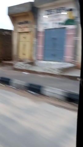 The video shows a blurred, moving scene from inside a vehicle. The view is out the side window. The environment appears to be a street with buildings on one side and a road on the other. The buildings are mostly simple, some appearing to be shops or small houses, with weathered facades and various colored doors and windows. Some of the buildings are in a state of disrepair, with open doorways and unkempt surroundings. There are patches of green grass and some debris on the ground in front of the buildings. The road has a curb with black and white markings. The sky is overcast. The overall impression is of a somewhat impoverished urban or semi-urban area.