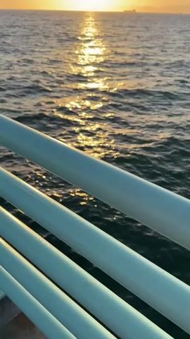 The video displays a scenic view of the ocean at sunset. The sun is a bright, warm orb positioned on the horizon, casting a shimmering golden path across the rippling water. The foreground is occupied by the light blue railing of a pier or balcony, which is angled diagonally across the frame, creating a sense of depth and perspective. The water appears calm with gentle waves. In the distance, a faint silhouette of land can be seen, along with a small ship or structure on the horizon. The sky transitions from a bright yellow near the sun to a softer orange and then a pale blue at the top of the frame. The overall ambiance is serene and peaceful.