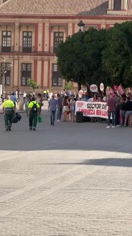 The media file depicts an outdoor scene in Sevilla, Spain, likely during the daytime under clear skies. The primary subjects are a group of people gathered in a plaza, holding a banner and some flags. The banner, prominently displayed, reads "SECTOR DE LIMPIEZA EN LUCHA" and features the logo "CCOO hábitat". This text indicates a protest or demonstration by workers in the cleaning sector, expressing their struggle or fight.

In the foreground, several individuals are walking across the paved plaza. One person is wearing a high-visibility green vest and carrying a green bag, possibly indicating their profession related to the cleaning sector. Behind this person, another individual in a similar vest is also walking. Further back, a larger group is congregated around the banner. They appear to be listening to someone or observing the display. Among the crowd, some individuals are holding round signs with the "CCOO hábitat" logo and what appear to be red flags. The people's expressions are not clearly discernible due to the distance, but the context of the banner suggests a serious or determined mood.

The setting is characterized by an impressive building with a red-tiled roof and ornate architectural details in the background, suggesting a historical or significant location. Large, mature trees with dense green foliage provide shade in the mid-ground. In the latter part of the media file, a grand cathedral or similar large, ancient religious structure with a tall bell tower is visible, with intricate stonework and arches. This architectural style strongly suggests a Spanish setting, reinforcing the "Sevilla, Spain" location context. The overall atmosphere appears to be one of a public gathering, possibly a rally or demonstration, taking place in a well-known urban square. The sunlight suggests it is likely late morning or afternoon.