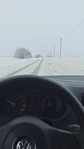 A dashboard camera records a car driving on a straight, snowy, rural road during the daytime. The road is partially cleared, with a single set of tire tracks visible. Snow covers the sides of the road, with sparse trees and utility poles lining the landscape. The dashboard shows the car's speedometer fluctuating around 90 km/h.  A digital display indicates the time is 17:16 and the outside temperature is 19 degrees Celsius.  Another car is visible ahead in the distance. A Volkswagen steering wheel is prominent in the foreground. Rap music plays in the background.