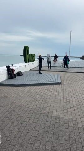 A person in black clothing and a yellow hat stands on a small grey wooden platform, playing a violin. Next to the platform is a white brick wall with a large green sign that says “OMAKS.” Behind the wall is a body of water with a clear horizon. Three other people are visible near the wall; one walking and two sitting on the wall. To the side of the platform are a violin case, a black speaker, and a small suitcase. The ground is paved with grey bricks. The sky is overcast.