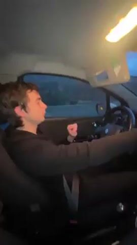 A young man is in the driver’s seat of a car, moving his arms rhythmically to music. He is wearing a black hoodie and pants. The car is traveling on a highway, with mountains visible in the distance. The interior light of the car is on. The driver looks at the camera briefly.