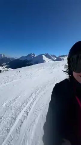 A person is skiing downhill in a snowy mountain environment, likely at a ski resort. The first-person perspective shows the skier wearing a dark helmet and goggles, with pink straps visible. The skier navigates the slope, casting a long shadow on the snow. Other skiers are visible further down the slope. Ski lifts are seen in the background, and the weather appears sunny.  The skier remains the primary focus throughout the video as they continue their descent.