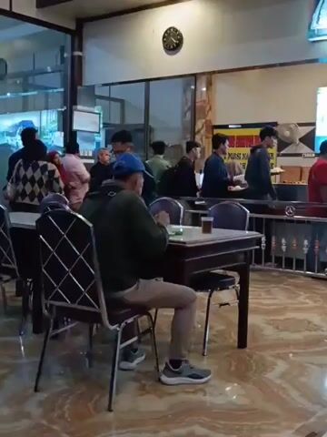 The video shows the interior of a restaurant with several people. In the foreground, a person in a green jacket and tan pants is seated at a table, eating. A cup of tea is on the table in front of them. The table is dark wood with a light-colored surface. The chair is dark with a silver frame. Further back, there is a counter with multiple people standing in line and being served. Above the counter are large screens displaying images, possibly food items or advertisements. A clock is visible on the wall. The flooring is tiled with a patterned design. People are dressed casually, and the overall atmosphere appears to be a busy dining establishment.