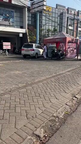 A white van is parked on a brick sidewalk next to a street. A person wearing a white shirt and dark pants is standing next to the open rear door of the van. There is a red tent set up on the sidewalk behind the van. Several motorcycles are parked under the tent. A building with large windows is visible behind the tent. There are signs on the building. The sky is clear and sunny.