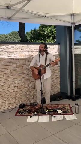A man with long dark hair and a beard, wearing a white shirt, light-colored pants, and sandals, stands under a white tent playing an acoustic guitar and singing into a microphone. He is positioned on a rug with various electronic devices and papers spread out around him. Behind him is a stone wall and some trees. The scene appears to be outdoors, possibly on a patio or deck.
