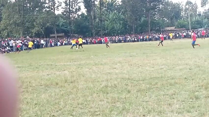 A wide shot shows a soccer game in progress on a dry grass field. A crowd of spectators stands behind a low fence or barrier, lining the edge of the field. The players are scattered across the field, with a cluster of players in red and yellow uniforms near the center, actively engaged in play. Several players in red uniforms are visible on the right side of the field, with one player in a yellow uniform further to the right. The background consists of trees and some simple buildings. The sky is overcast. The camera appears to be held by someone at ground level, with a thumb visible in the lower left corner of the frame. The video quality is blurry and grainy.