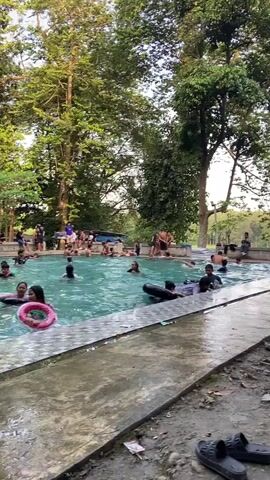 The video shows a crowded outdoor swimming pool on a sunny day. Numerous people, including adults and children, are in the water, swimming, splashing, and playing with inflatable toys like tubes and rings. Some individuals are sitting on the edge of the pool, while others are standing or wading in the shallower areas. The pool is surrounded by lush green trees and vegetation, indicating a natural or semi-natural setting. There are also some man-made structures visible in the background, such as umbrellas and what appear to be stalls or seating areas. The overall atmosphere appears to be one of recreation and enjoyment.