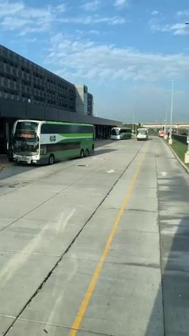A wide shot captures a bus station with a modern building and several buses. In the foreground, a green and white double-decker bus is parked on the left. Ahead, a road marked with yellow and white lines curves to the right. Several other buses, including a smaller white one and another green and white bus, are visible at the station in the middle ground. People can be seen standing and walking near the station entrance. The sky above is blue with scattered clouds. The overall scene suggests a functional transportation hub on a clear day.