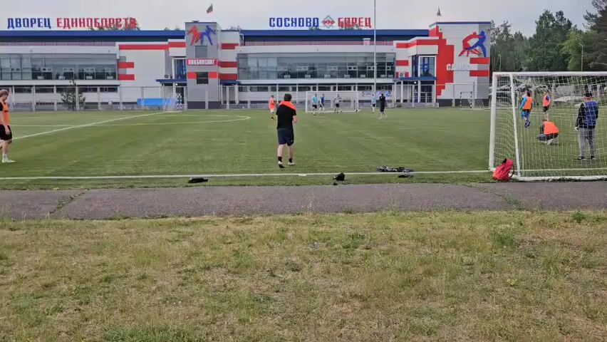 The video shows an outdoor soccer field with a group of people, mostly young males, engaged in a game or practice. The field is green and well-maintained, with white boundary lines. A soccer goal with a net is visible on the right side of the frame. In the background, there is a large, modern building with a sign that reads "ДВОРЕЦ ЕДИНОБОРСТВ" and "СОСНОВО БЕРЁЗ". The building appears to be a sports complex. The sky is overcast, suggesting a cloudy day. People are scattered across the field, some running, some standing, and some interacting with a soccer ball. One person in the foreground is facing away from the camera, wearing an orange shirt and dark shorts. Another person in an orange shirt is running to the left. On the right side, near the goal, a few people are gathered, one of whom is crouching. The scene appears to be a casual sports activity taking place at a dedicated facility.