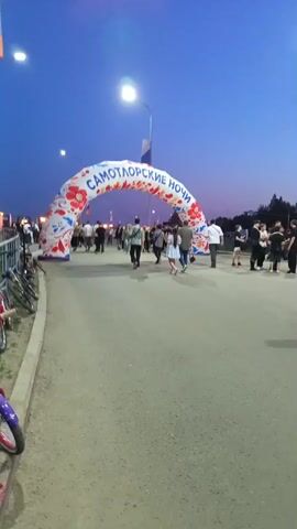The video shows a wide shot of an outdoor scene at dusk. In the foreground, a paved path leads toward a large, inflatable archway. The archway is white with red and blue floral designs and has Russian text that reads "САМОТЛОРСКИЕ НОЧИ" (Samotlor Nights). Several people are walking through or standing near the archway. The lighting suggests it is evening, with streetlights visible and a deep blue sky. To the left of the path, a row of bicycles is parked along a fence. The overall atmosphere appears to be that of an event or festival.