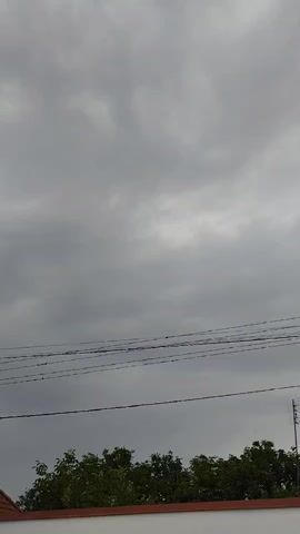The video shows a cloudy sky with a few power lines running horizontally across the frame. Below the power lines, there are trees and the top of a building with a red trim. The overall impression is of an overcast day, possibly before or after rain. There are no people or any discernible actions taking place. The scene is static and captures a brief moment in time.
