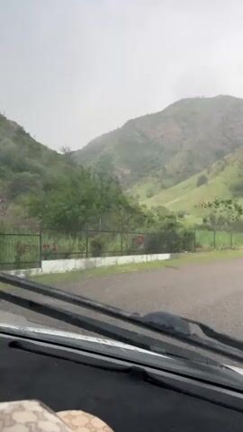 A view from inside a vehicle shows a mountainous landscape with green hills and scattered trees under a cloudy sky. Raindrops are visible on the windshield, and a windshield wiper moves across the frame. The road in front is wet. A fence runs along the side of the road, separating it from the fields and hills.