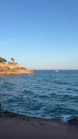 The video shows a view of the ocean on a clear day with a sailboat in the distance. In the foreground, there are rocks and waves crashing against them. To the left, there is a rocky outcrop with some trees on top. The sky is a clear blue. The sailboat appears to be white with a single sail. The water is a deep blue and appears choppy.
