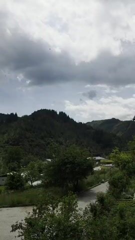 The video presents a panoramic view of a mountainous landscape under a heavily clouded sky. The scene begins with a wide shot dominated by verdant, tree-covered mountains stretching across the horizon. A paved road winds through the middle ground, bordered by lush green foliage. Faint dark specks, possibly birds, are visible against the overcast sky.

As the camera slowly pans from left to right, a cluster of buildings comes into view on the right side of the frame. The most prominent structure is a house with weathered, light-colored walls and a distinctive brown tiled roof, characteristic of traditional architecture in the region. A solar water heater is visible on its roof, and a second, similar building is partially obscured behind it.

The foreground features a well-tended garden area with various plants and a white tarp covering part of the ground, indicating cultivation. A satellite dish is partially visible in the bottom right corner, suggesting the vantage point is from a nearby elevated position. The sky remains consistently overcast throughout the video, creating a soft, diffused light. No specific human activity is depicted. The overall impression is one of a serene, rural setting nestled within the mountains. The location is Rudozem, Bulgaria. The audio accompanying the video appears to be the Islamic call to prayer (Adhan). No visible text is present in the video.