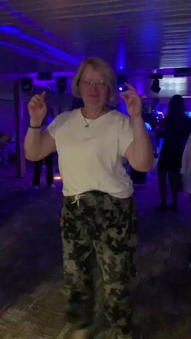 A woman with blonde hair, wearing a white t-shirt and camouflage pants, dances in a dimly lit room with purple and blue lights. She is facing the camera and makes various hand gestures. There are other people in the background, some dancing and some standing. The floor has glowing green patterns on it.
