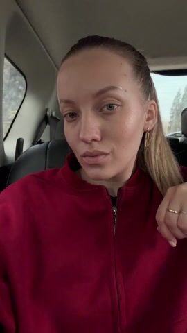 The video shows a fair-skinned woman with light blue eyes sitting in the back seat of a car. She is wearing a red zip-up jacket and gold earrings. Her light brown hair is pulled back in a ponytail. She glances at the camera, then rests her face on her hand. The car's interior is black. A window and the back windshield are visible behind her. The weather outside appears overcast. Music plays in the background.