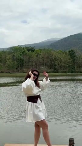 A woman stands on a wooden dock at the edge of a lake, with lush green trees and mountains in the background under an overcast sky. She wears a short white dress cinched at the waist with a wide, dark brown belt, complemented by a heart-shaped red purse and sunglasses. She is smiling and strikes various poses, extending her arms out to the sides and waving at the camera. At one point, she lifts one leg up in the air. She also turns around to face the scenery while still waving.