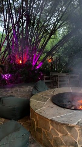 The video shows an outdoor seating area with a fire pit at night. A circular fire pit is made of stone, with a metal bowl holding a fire burning inside. Several beanbag chairs are placed around the fire pit. In the background, bamboo trees are lit with purple and red lights. A table and chairs are also visible further back in the outdoor space. Smoke from the fire drifts through the air.