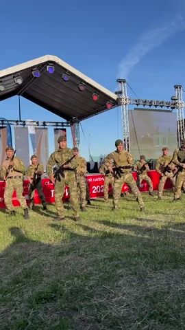 A group of soldiers perform a choreographed dance on a grassy field in front of a stage. The soldiers are dressed in camouflage uniforms with green berets. They wield rifles and move in sync to music. The stage behind them has a screen displaying colorful graphics. The sky is clear and blue. There are banners and flags visible on the stage and in the background, with the word "СПОРТИВНО-ТУРИСТСКИЙ ЛАГЕРЬ ПФО 2026" in Russian.
