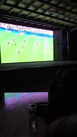 A crowd of people are gathered outdoors, seated in chairs and on the ground, watching a large screen that displays a soccer match. The screen is brightly lit, and the surrounding area is dark, suggesting it is nighttime. The people appear to be casually dressed and some are holding drinks. The atmosphere seems relaxed, and many are looking towards the screen, engaged in watching the game.