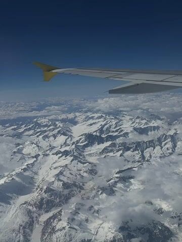 An aerial view from an airplane shows a vast, snow-covered mountain range under a clear blue sky. The white wing of the aircraft, with a yellow tip, extends into the upper right of the frame, partially obscuring the sky. Below the wing, numerous jagged mountain peaks are blanketed in white snow, with some visible rock faces. Clouds are scattered among the lower parts of the mountains and fill some valleys, creating a sense of depth. The scene suggests a bright, clear day, with sunlight illuminating the pristine, snowy landscape. The location context provided indicates this is likely the Alps near Göschenen, Switzerland. No people or text are visible.