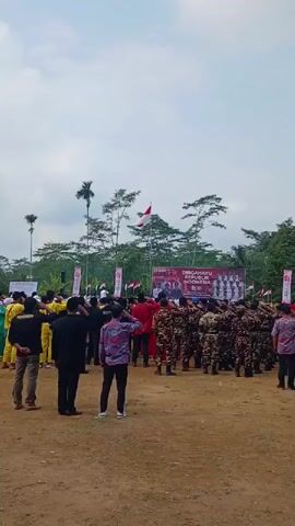 A crowd of people stand in an outdoor field, facing a stage with banners. Several individuals in military camouflage uniforms stand at attention and salute. Other people in civilian attire, including some in yellow uniforms, also face the stage. The Indonesian flag flies from a pole behind the stage. The sky is overcast.