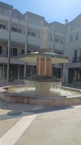 The video pans across a fountain in a courtyard. The fountain is multi-tiered and made of stone. Water cascades from the upper tiers into the basin below. Behind the fountain is a modern building with several floors, balconies, and shops on the ground level. The sky is clear and blue, with the sun shining brightly. The courtyard is paved with light-colored stone tiles. The overall scene is tranquil and picturesque.