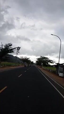 The video shows a wide, paved road from the perspective of someone traveling on it. The road has a yellow line down the center and a white line on the right edge. There are trees on both sides of the road, some of which are quite tall. There are also some small buildings or structures along the right side of the road. The sky is overcast with heavy clouds. Several vehicles are visible on the road, including motorcycles and what appears to be a truck or van. Some people are also seen walking along the side of the road or riding on the vehicles. The overall scene depicts a rural or semi-rural setting on a cloudy day.