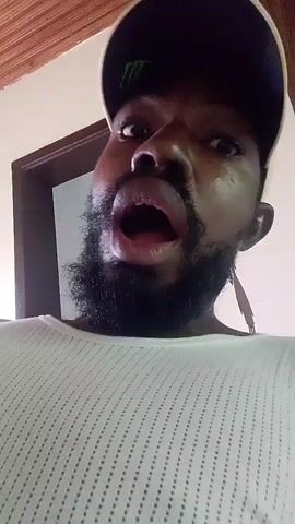 A close-up, point-of-view shot shows a Black person wearing a white, ribbed shirt and a baseball cap. The person has a beard and is speaking with their mouth open, revealing their teeth and tongue. The background shows a wooden ceiling and a door frame.