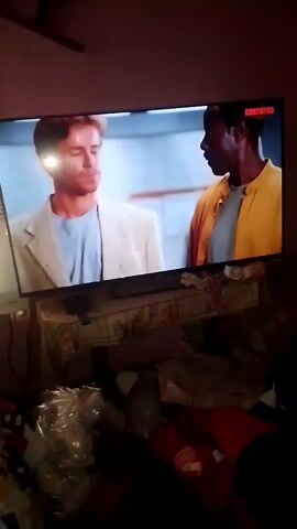 The video shows a television screen displaying a scene from a movie. On the left side of the screen, a man with blonde hair and a light-colored jacket is speaking. On the right side of the screen, a Black man in a yellow jacket is looking at him. The screen has a logo in the top right corner that reads "CINEMATCH". Below the television, a table is visible, covered with a patterned cloth. On the table, there are various items, including what appear to be plastic bags and a red garment. The lighting in the room appears dim. The scene transitions to show a different man with graying hair, wearing a light blue shirt and a tie, speaking to the camera. He is in an office-like setting with framed pictures on the wall behind him. The video then returns to the scene with the blonde man and the Black man.