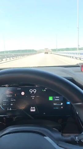 The video shows the perspective of someone driving a car on a highway bridge. The bridge is over water and has metal guardrails. A truck is visible in the distance on the highway ahead. The side view mirror shows the empty highway behind the car. The sky is blue with some wispy clouds.