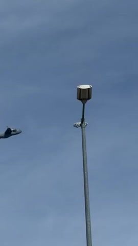 A gray Airbus A400M Atlas military transport aircraft flies in the sky, passing behind a gray metal light pole with a white box-shaped light fixture at the top. The sky is light blue and mostly clear with scattered clouds. The airplane appears to be in flight.