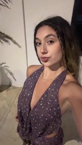 A young woman with long, wavy brown hair and a deep purple sequined halter top with a plunging neckline poses for a video. She is standing outdoors against a plain wall with a hint of greenery to her left. Her hair is partly braided and loose. She playfully pouts her lips and touches her hair and neck. The video is shot from a close-up perspective, and the lighting is dim, suggesting it is nighttime or an indoor setting with low light. The woman's movements are quick and fluid, indicating she is likely recording a selfie video or a short clip for social media.