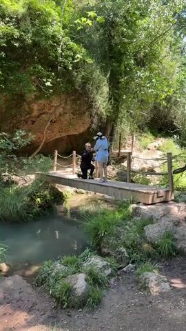 A man is kneeling on a wooden bridge over a stream. He is wearing a dark jacket and black pants. He is looking up at a woman who is standing in front of him. The woman is wearing a light blue dress and a white hat. The bridge is surrounded by trees and rocks. There are ropes on the sides of the bridge for support. The stream is clear and there is green grass on the banks.