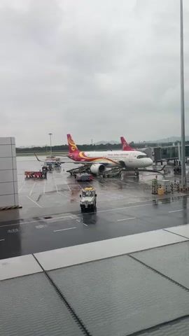 A wide shot shows an airplane parked at an airport gate on a cloudy day. The plane, which is red and white with a yellow tail, has "Hainan Airlines" written on its side in English and Chinese. Several ground vehicles, including a baggage tug, are parked near the plane, and some airport personnel can be seen around the aircraft. The tarmac is wet, indicating that it has recently rained. The scene appears to be a typical airport operations setting.