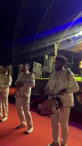 A band of three musicians performs on a red carpeted stage. The musician on the left plays a trumpet. The middle musician plays a trumpet. The musician on the right plays a drum and has drumsticks. They are all wearing white suits with gold trim and sunglasses. The background is dark with stage lighting.