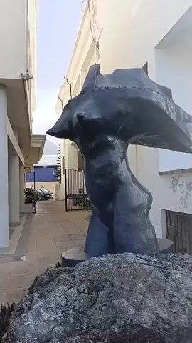 A dark, abstract sculpture resembling a torso and head stands on a large rock in an outdoor walkway. The sculpture is positioned to the right of the frame. The walkway is lined with buildings on both sides. The building on the left has white columns and a sign that reads "53". The building on the right appears to be a lighter color. In the background, a car is visible in a parking area. The sky is clear and bright.